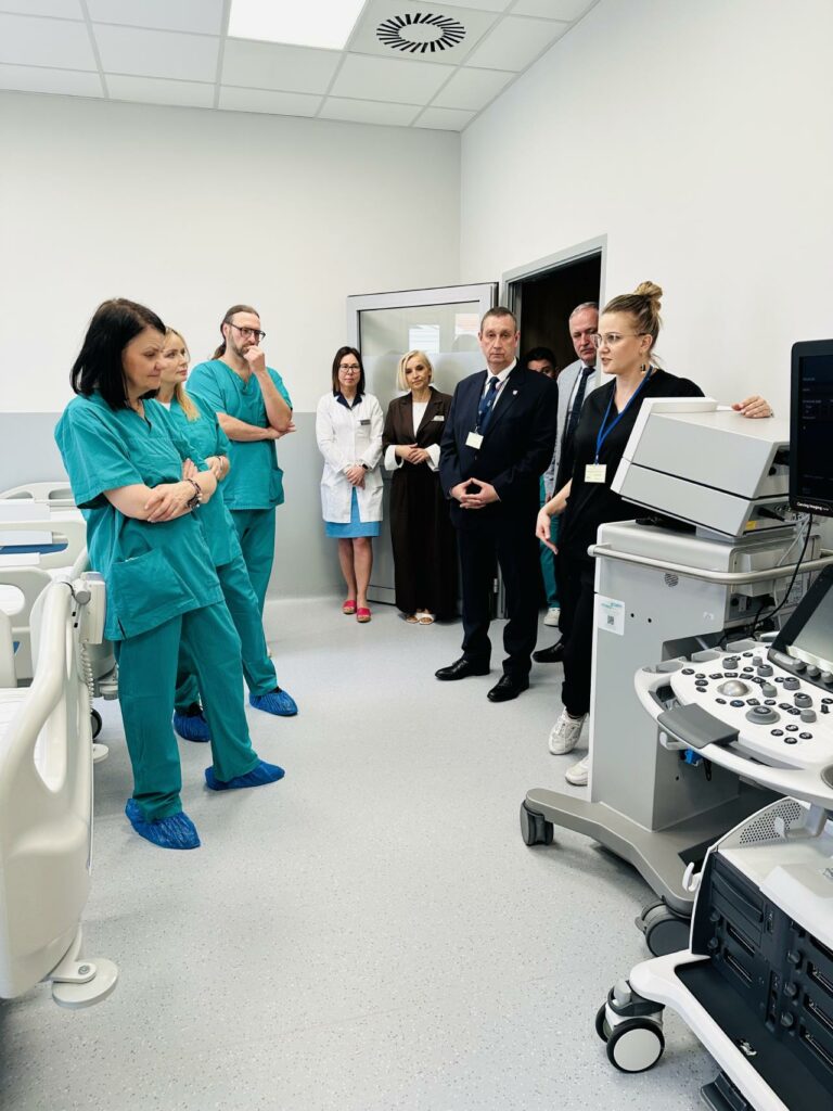 Zdjęcie przedstawia grupę osób zgromadzonych w nowoczesnej sali szpitalnej podczas prezentacji sprzętu medycznego