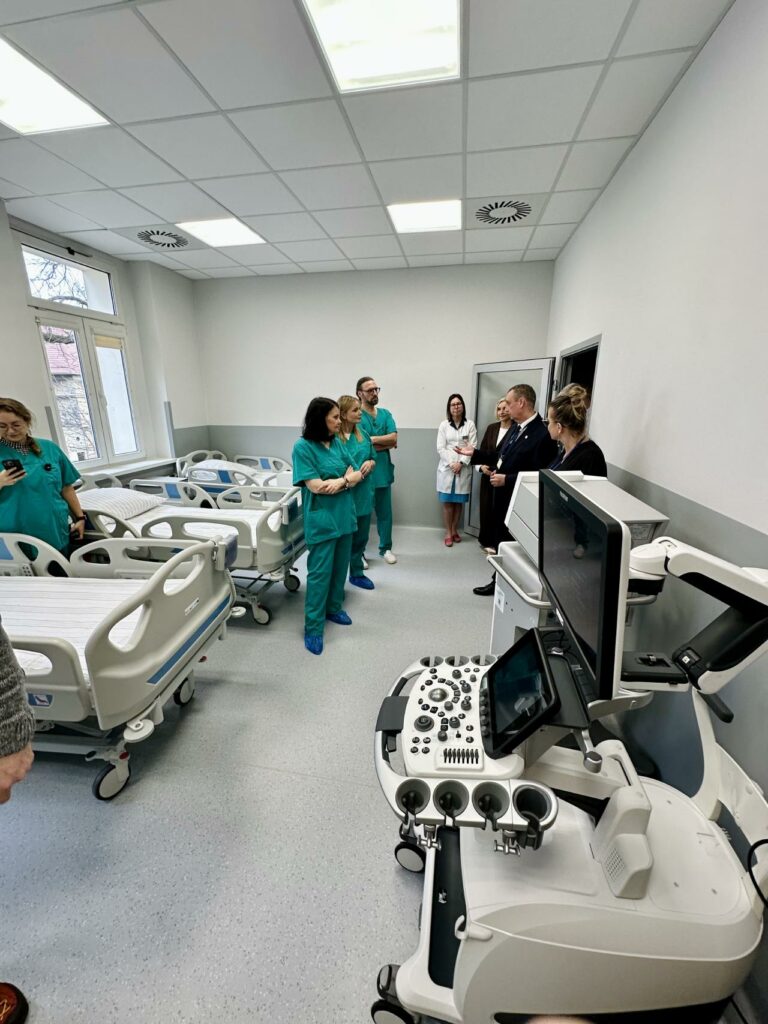 Zdjęcie przedstawia grupę osób zgromadzonych w nowoczesnej sali szpitalnej podczas prezentacji sprzętu medycznego