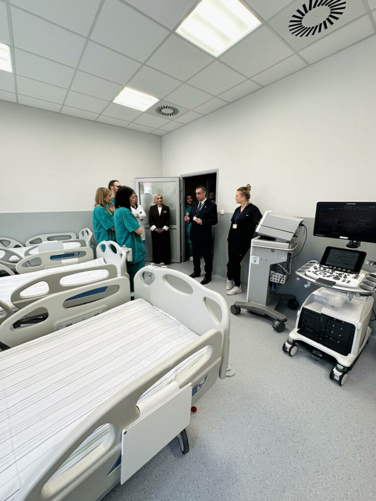 Zdjęcie przedstawia grupę osób zgromadzonych w nowoczesnej sali szpitalnej podczas prezentacji sprzętu medycznego